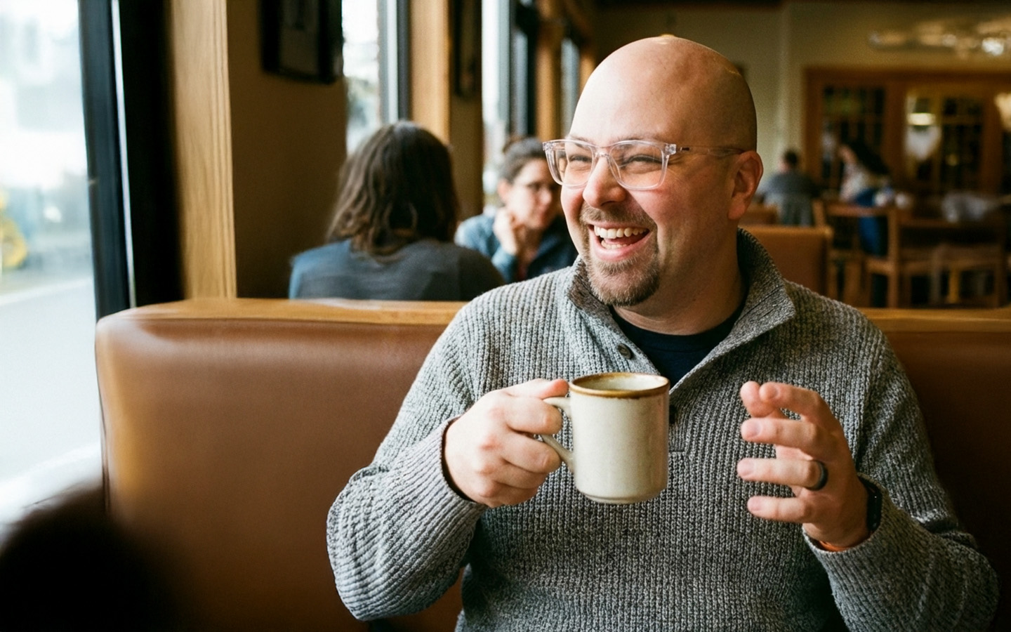 Simon smiling widely while sitting in a coffee shop with a cup of coffee in his hand.